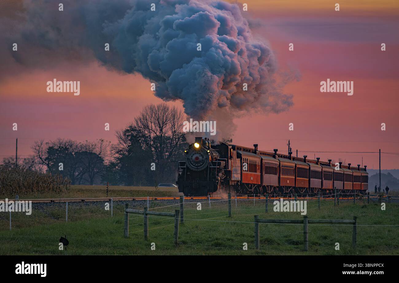 Una vista di un treno passeggeri a vapore antico che si avvicina all'alba con una testa piena di vapore e fumo che viaggia attraverso le terre Foto Stock
