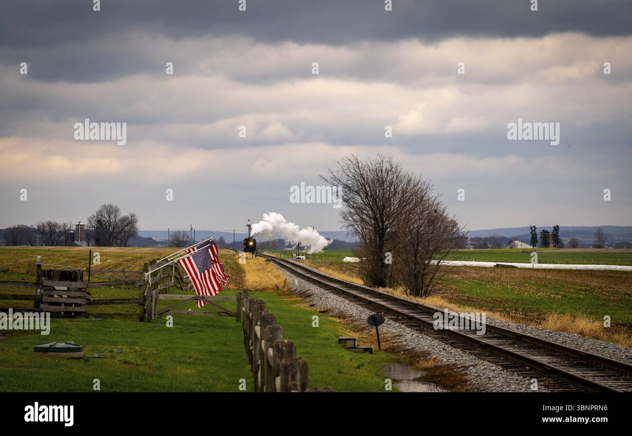 Un motore di vapore e le vetture restaurate d'antiquariato si avvicinano a Thru Corn Fields con le bandiere americane che fodera la pista in un giorno di sole Foto Stock