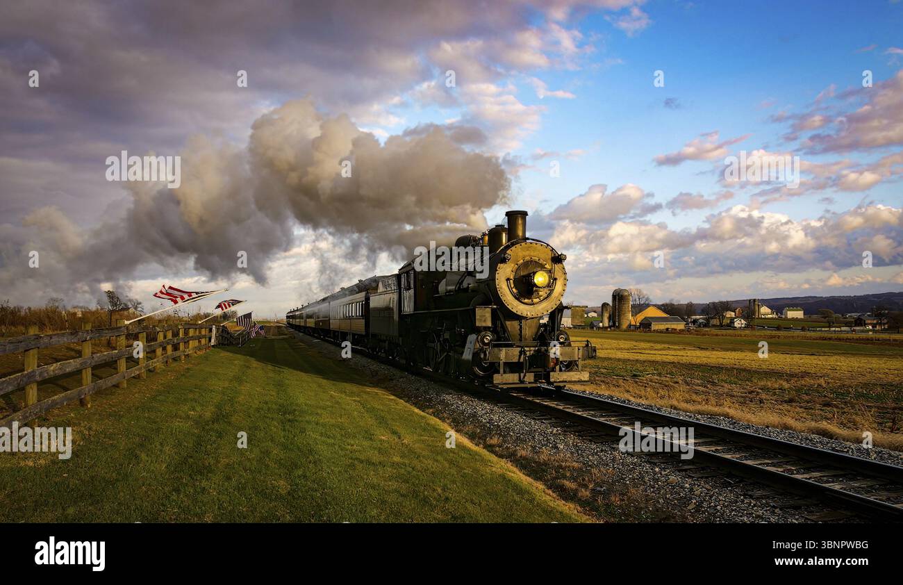 Una veduta di un treno passeggeri a vapore classico che si avvicina, con le bandiere Americane attaccate ad un recinto in un giorno di autunno soleggiato Foto Stock Una veduta di un treno passeggeri a vapore classico che si avvicina, con le bandiere Americane attaccate ad un recinto in un giorno di autunno soleggiato Foto Stock