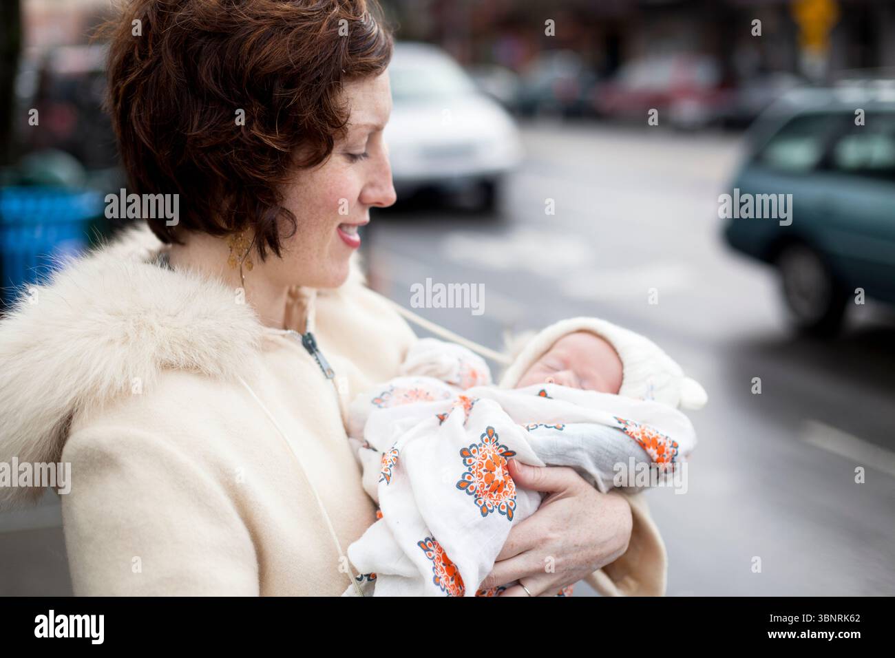 Una donna con i capelli rossi in un cappotto bianco vintage rifinito in pelliccia tiene un neonato ondulato tra le braccia su un marciapiede di una città. Foto Stock