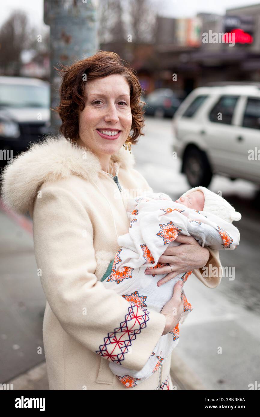 Una donna con i capelli rossi in un cappotto bianco vintage rifinito in pelliccia tiene un neonato ondulato tra le braccia su un marciapiede di una città. Foto Stock
