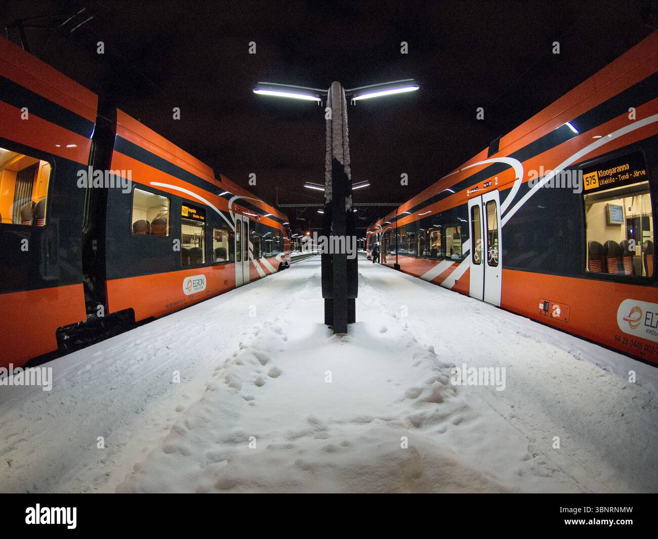 Moderni treni passeggeri arancioni sulla piattaforma ferroviaria innevata di notte, Estonia. Treno Stadler Foto Stock