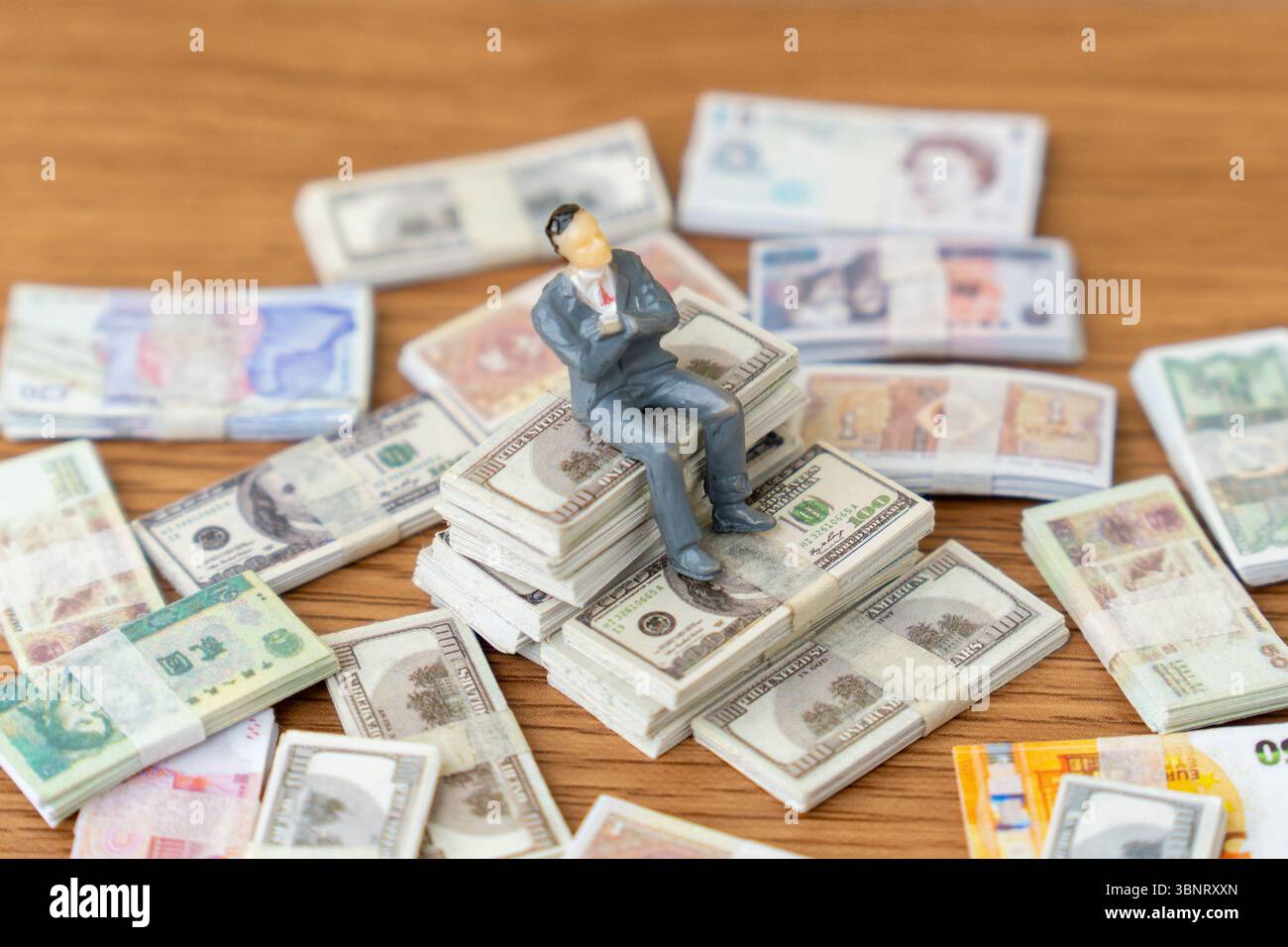 Un uomo d'affari in miniatura si trova in cima a una grande pila di banconote in dollari USA, circondato da altre valute internazionali. Ricchezza, successo finanziario Foto Stock