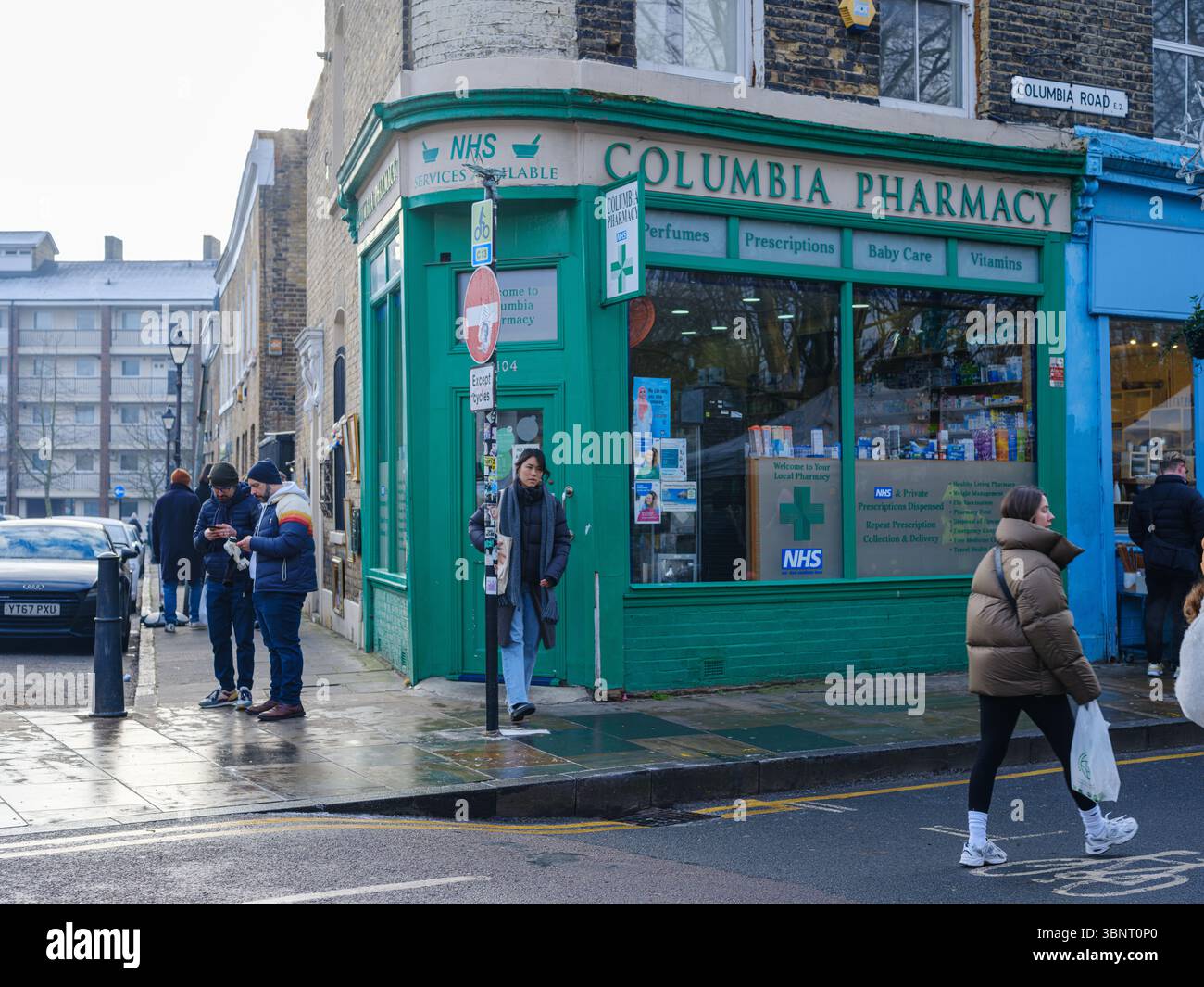 Londra, Regno Unito, 27 dicembre 2025. I pedoni passano davanti a una farmacia locale all'angolo di Columbia Road, con marciapiedi bagnati che riflettono la vita quotidiana della città Foto Stock