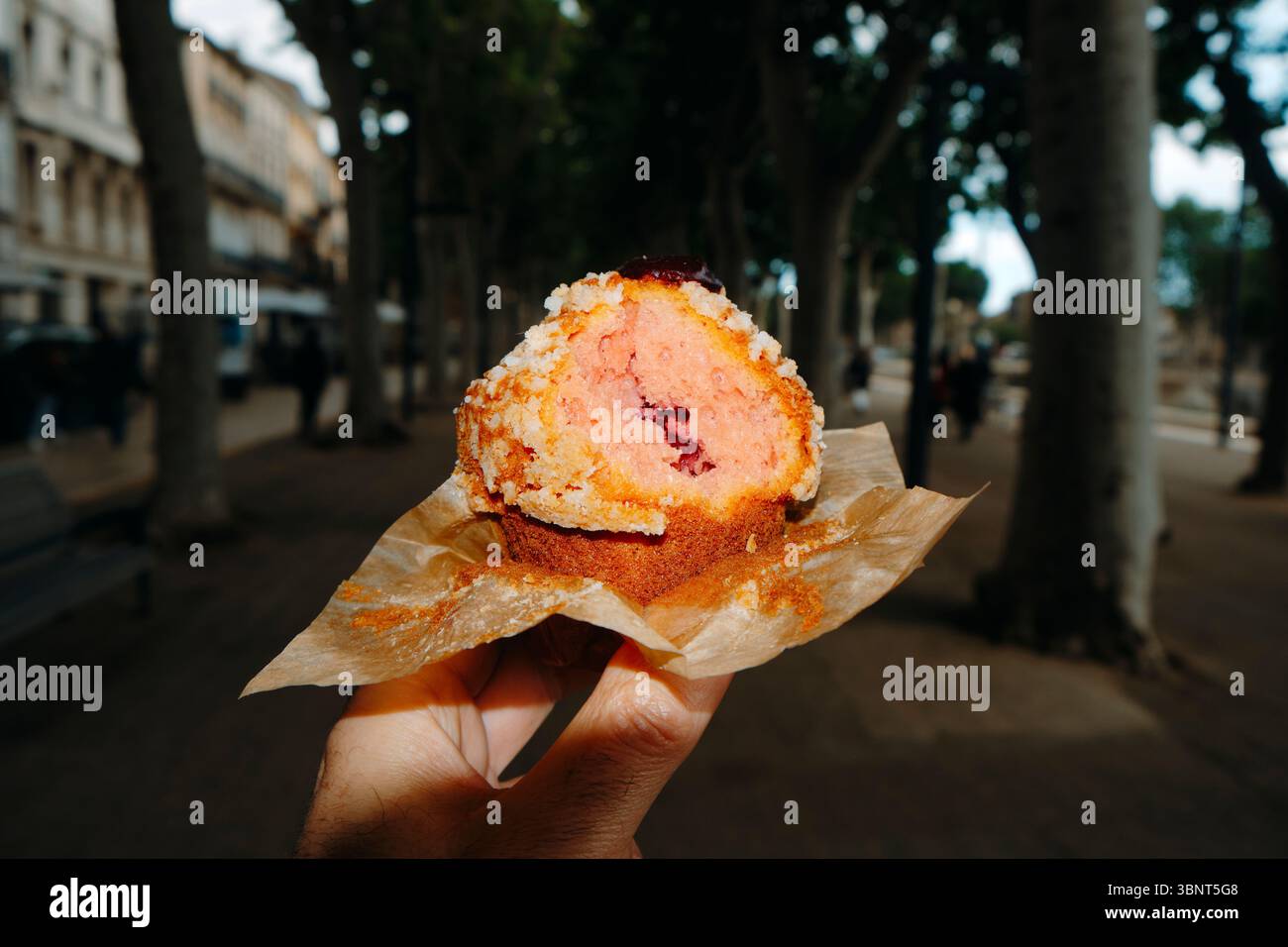 primo piano di un uomo che tiene in mano un muffin morso ripieno di marmellata di fragole, appena sfornato in una panetteria locale, in piedi su una passeggiata alberata Foto Stock