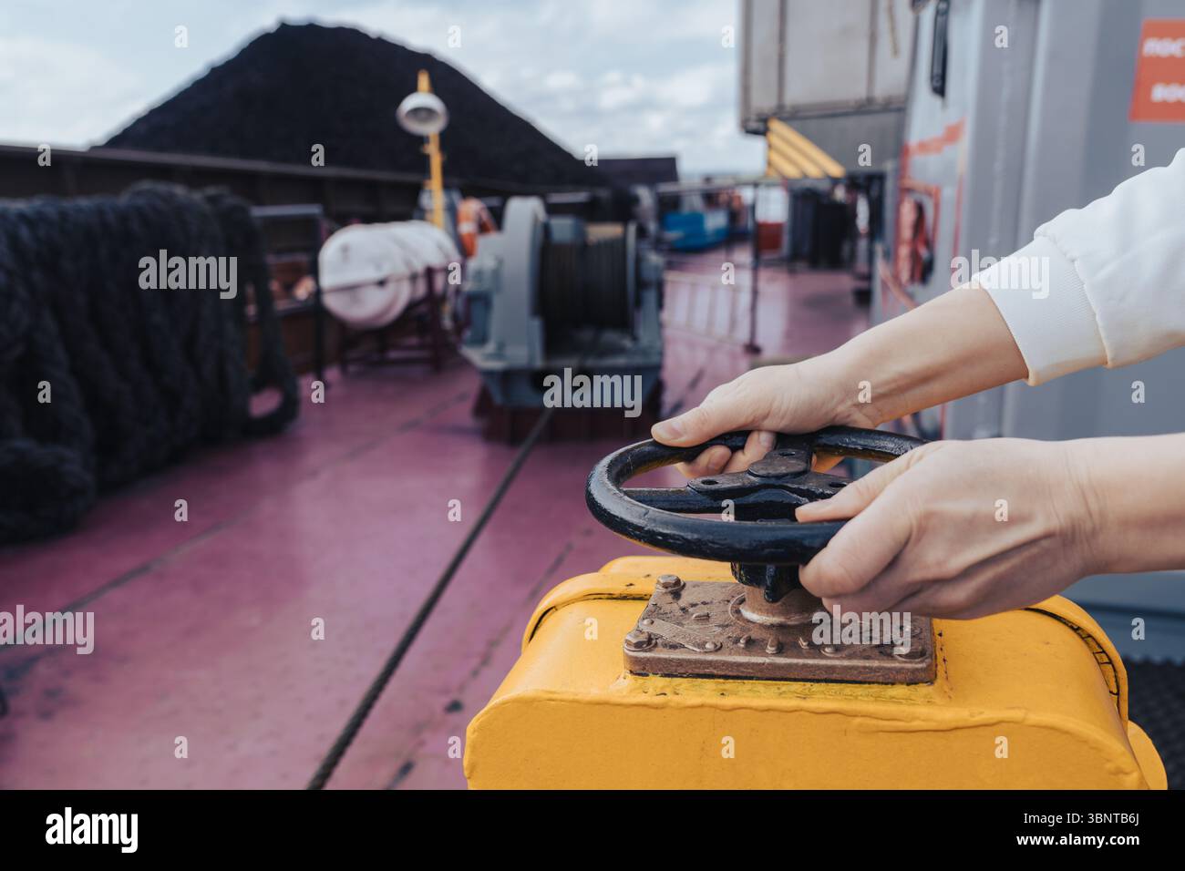 Un membro dell'equipaggio utilizza una valvola di controllo sul ponte di una nave da carico, gestendo con competenza le operazioni durante il carico di carbone in un porto vivace. La ripresa della scena Foto Stock