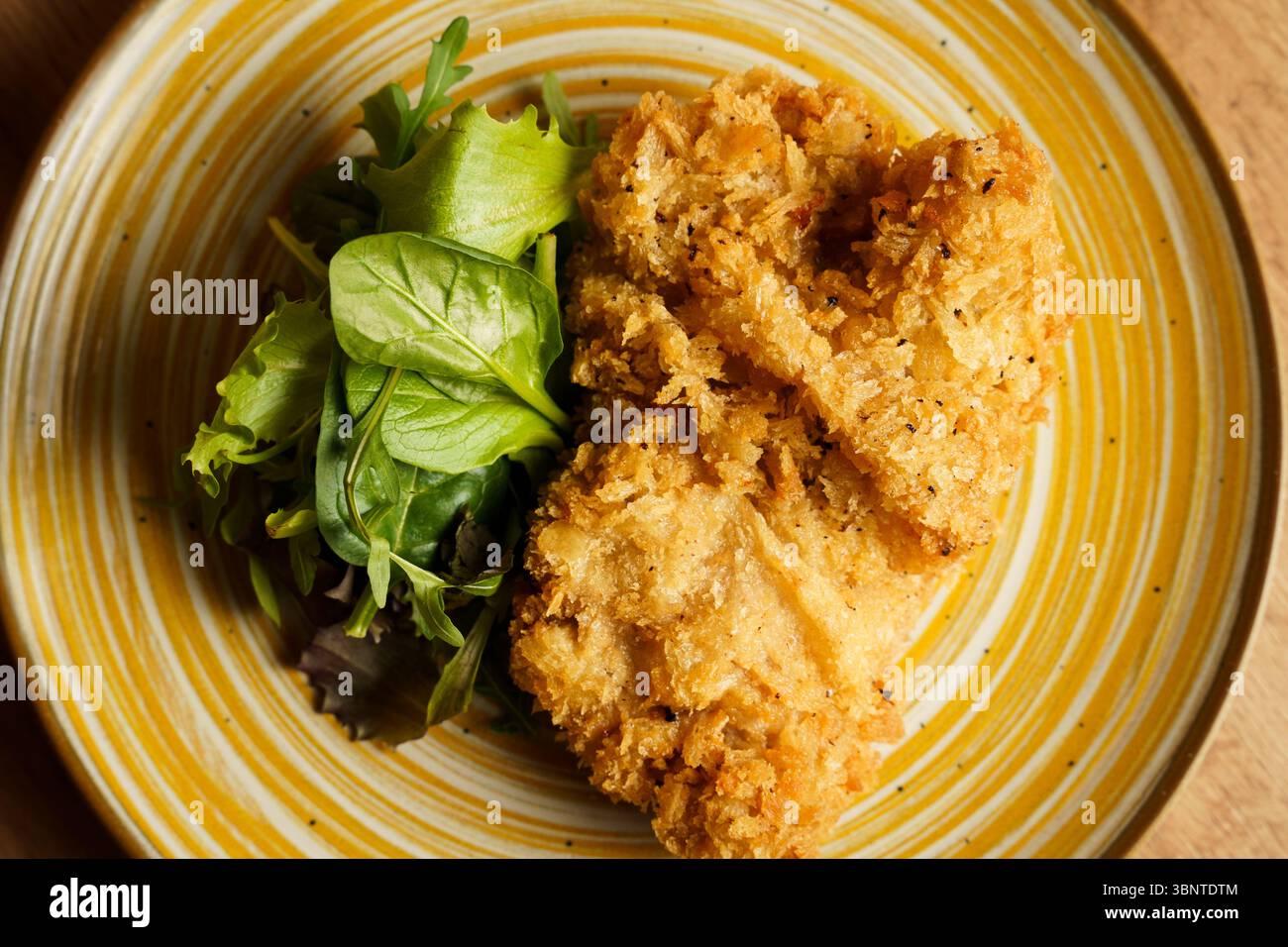 Filetti di pollo fritti dorati serviti con un lato di vivaci insalate miste su un piatto a strisce gialle. Il piatto contrasta la consistenza croccante wi Foto Stock