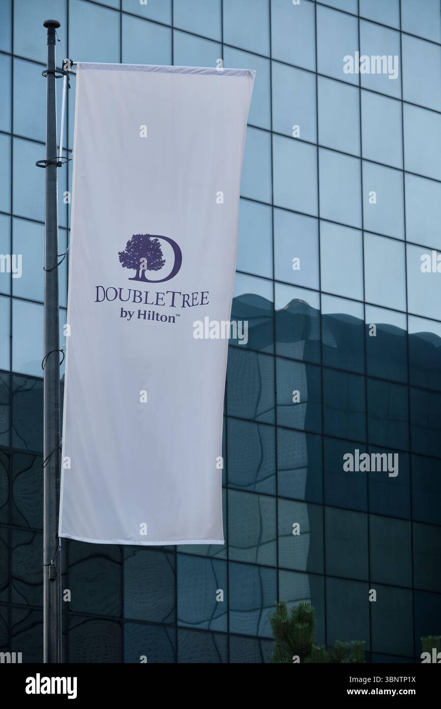 Lodz, Polonia - 2 luglio 2025: Verticale bianco DoubleTree sulla bandiera Hilton che sventola su un palo di fronte alla facciata di un edificio moderno. Foto Stock