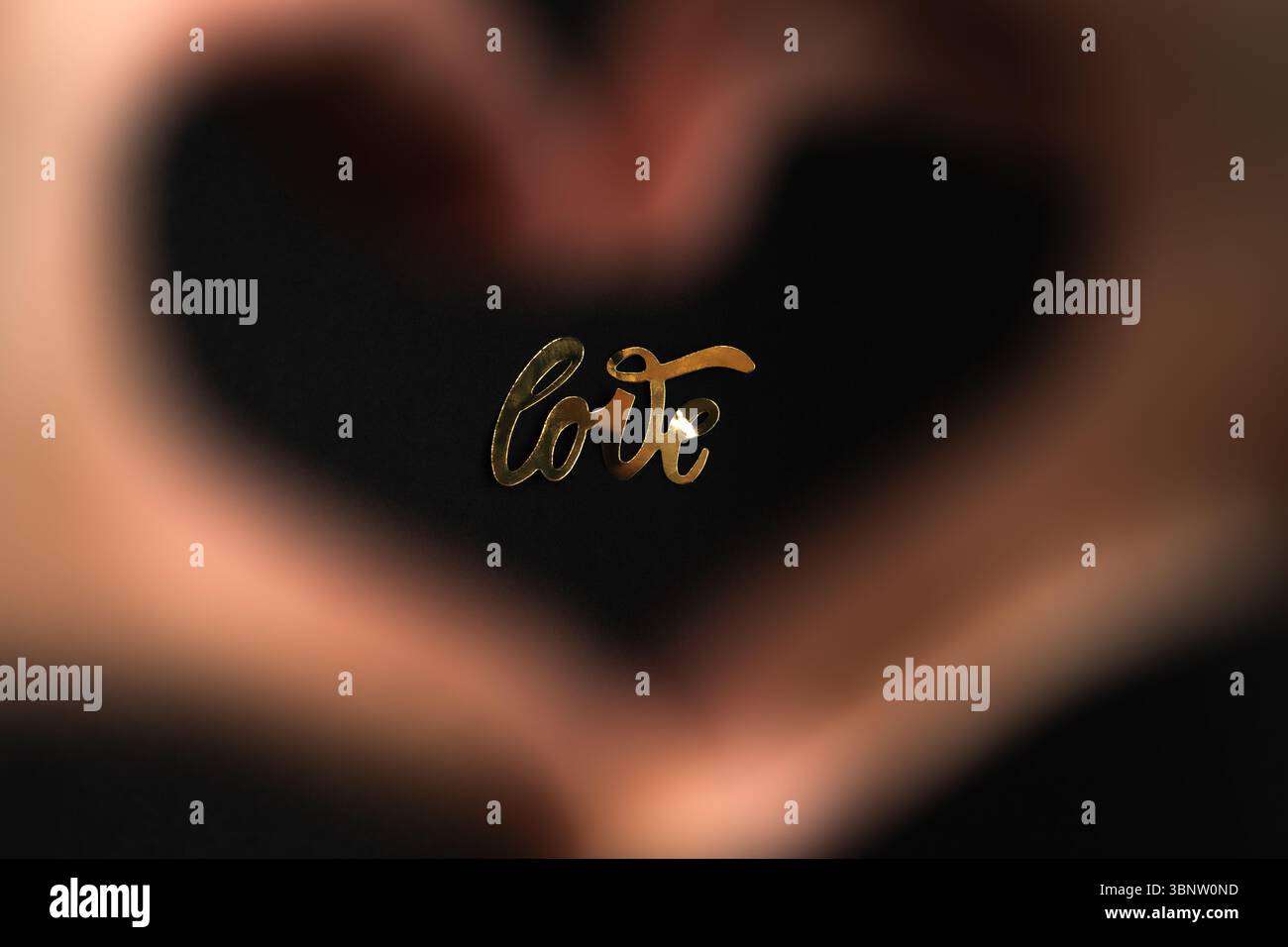 Amore. La parola "amore" brilla di lettere dorate, viste attraverso una forma a cuore fatta con le mani. Una fotografia romantica e creativa in primo piano. Foto Stock