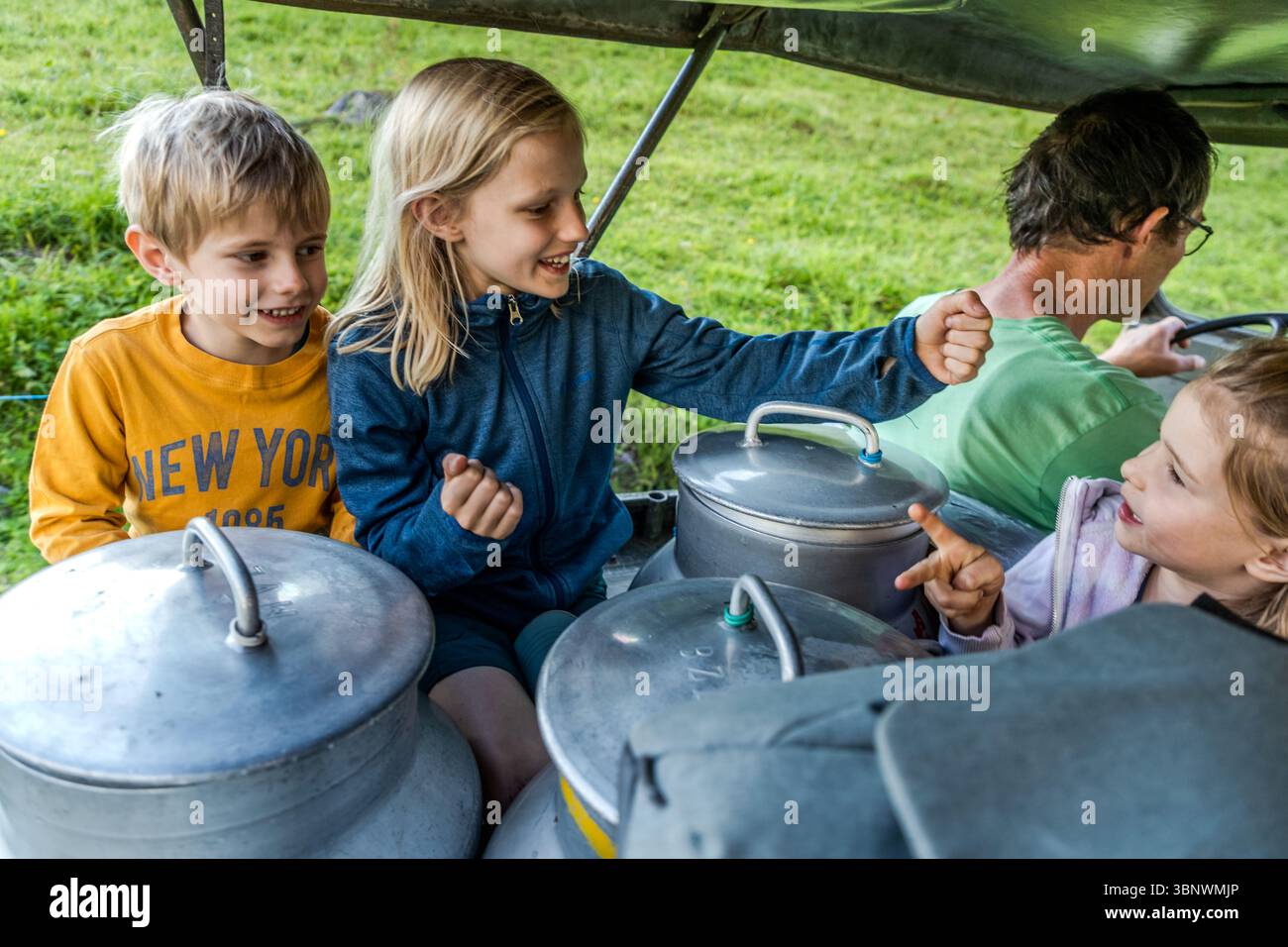 Tre bambini in vacanza in fattoria ridono e si scambiano un high-five sotto la capote della jeep, circondati da botti di latte sulla via del ritorno a Hobiel Alp. Ruedi Zurfluh guida il veicolo attraverso la valle di Engelberg vicino al passo di Surenen. Attinghausen, Cantone di Uri, Svizzera. Foto Stock