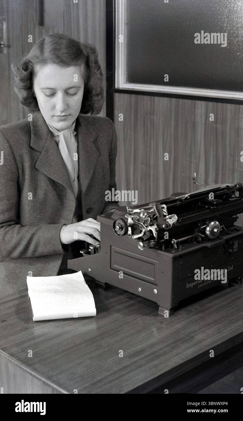 Anni '1940, storica ed elegante giovane signora, abito in lana e scolletto, una segretaria che usa una macchina da scrivere, realizzata dalla Imperial Typewriter Co di Leicester, Inghilterra, Regno Unito, scrivendo una lettera da un taccuino con stecche sulla scrivania. Foto Stock