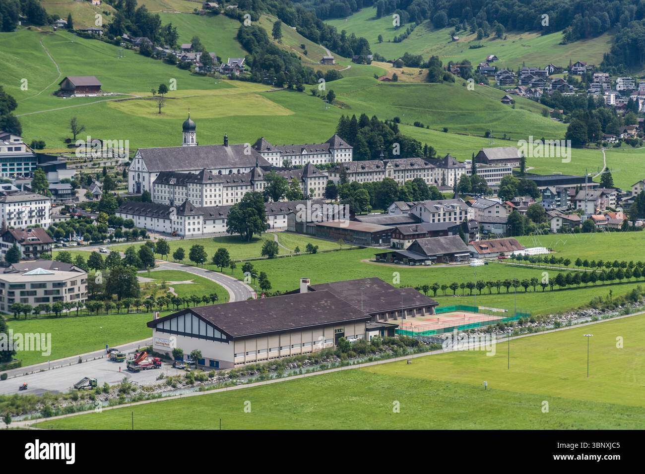 L'abbazia di Engelberg, fondata nel 1120, è una delle più antiche abbazie benedettine della Svizzera. Il complesso dell'abbazia è uno dei più importanti complessi barocchi della Svizzera centrale. Una vista panoramica mostra il monastero benedettino di Engelberg, circondato da prati verdi e il villaggio, incastonato nel pittoresco paesaggio della valle. Un complesso sportivo con campi da tennis e un fiume possono essere visti in primo piano. Engelberg, Canton Obwalden, Svizzera (CH), Europa Foto Stock