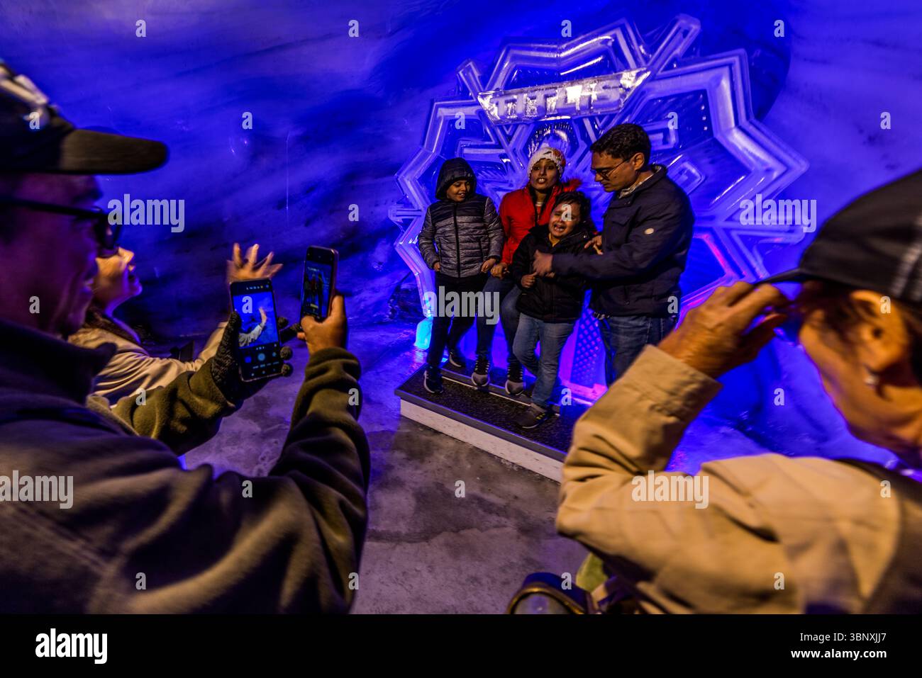 La grotta del ghiacciaio del Titlis è bagnata da un mistico crepuscolo blu turchese e le pareti sono coperte da milioni di scintillanti e antichi cristalli di ghiaccio che risalgono a 5.000 anni fa. La temperatura nella grotta rimane costantemente ghiacciata, meno 1,5 gradi Celsius. I visitatori, tra cui una famiglia, posano e scattano foto di se stessi nell'impressionante grotta di ghiaccio blu sul Titlis a Engelberg, Obvaldo, Svizzera. La scultura di ghiaccio "TITLIS" funge da sfondo suggestivo per le foto ricordo, mentre altri ospiti catturano l'atmosfera unica con i loro smartphone Foto Stock