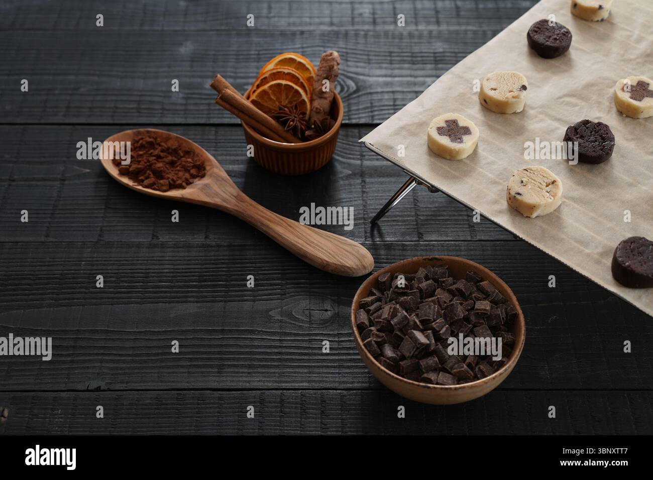 Primo piano dell'impasto per biscotti su una griglia da forno con cucchiaio di legno di cacao su un asciugamano da cucina, ciotola con gocce di cioccolato e misuratori in metallo vista frontale Foto Stock