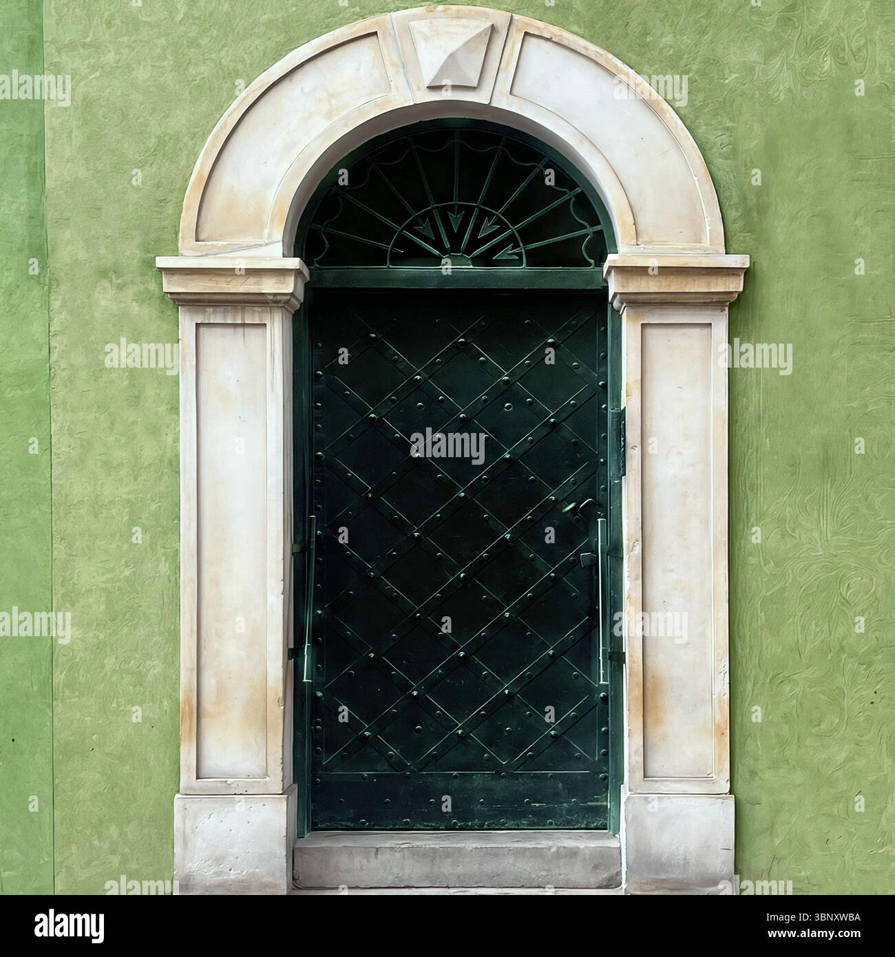 Una porta verde scuro rivestita in ferro con specchio di poppa soleggiato nella città vecchia di Varsavia si erge incorniciata da un arco in pietra neoclassico e pareti in gesso di olivo testurizzato. Foto Stock