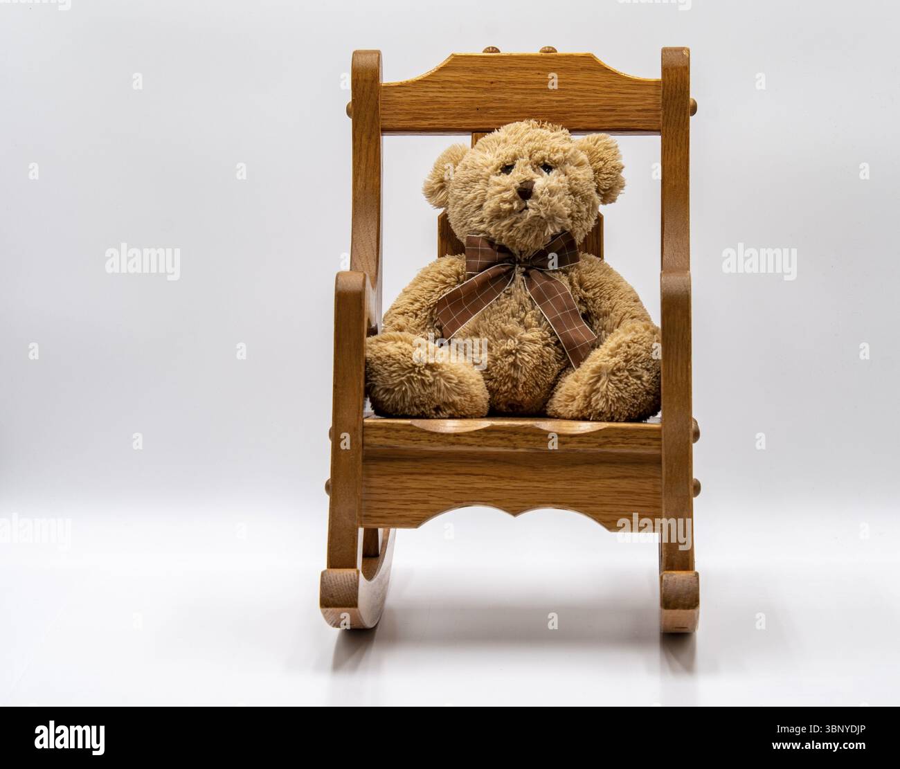 Un soffice orsacchiotto con nastro marrone siede comodamente su una sedia a dondolo in legno collocata in uno spazio interno pulito e minimalista. L'atmosfera generale lo è Foto Stock