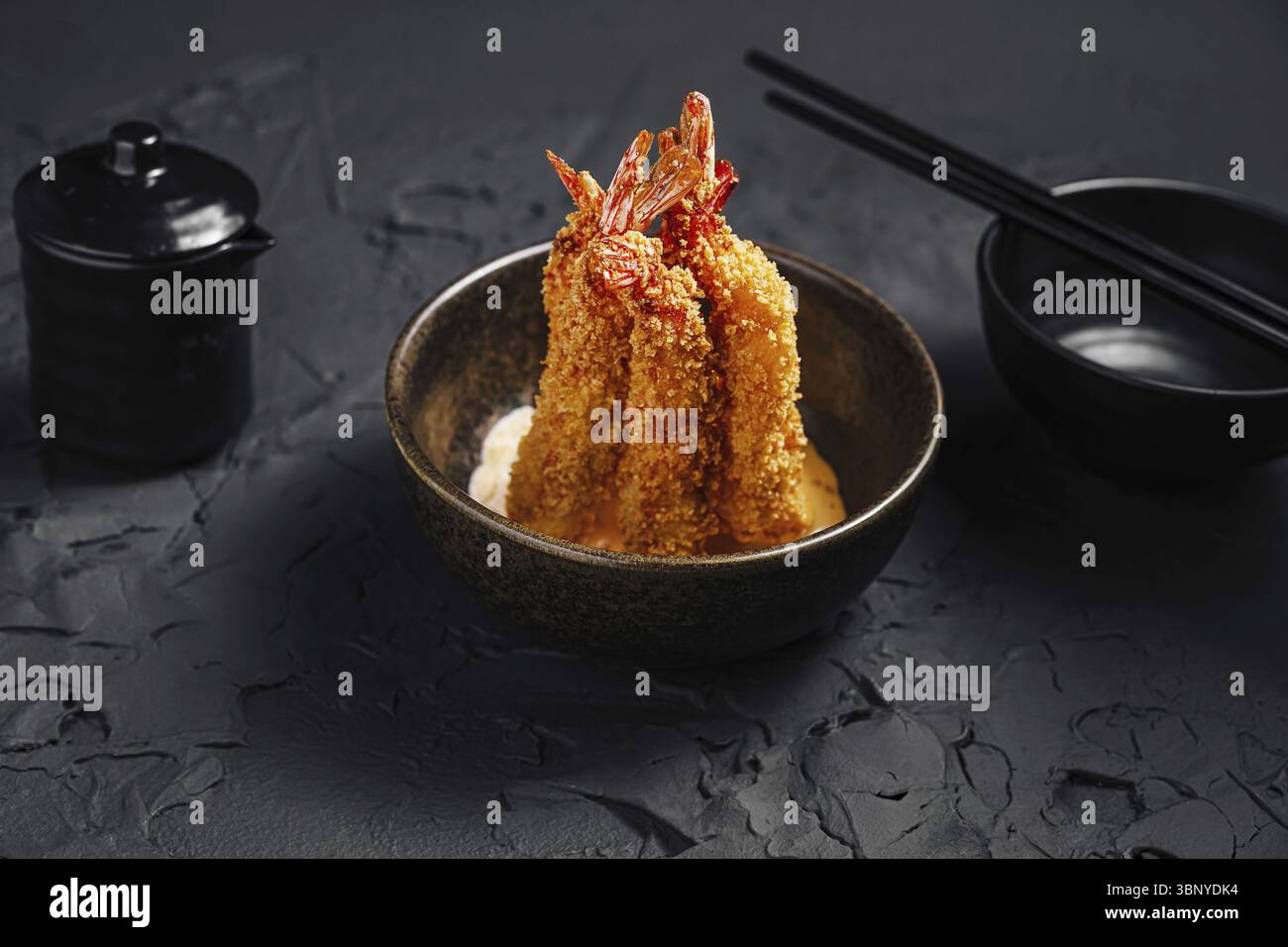 Cibo natura morta, gamberi impanati croccanti dorati sono elegantemente disposti in una ciotola scura, abbinati a condimenti e bacchette su una superficie nera testurizzata Foto Stock