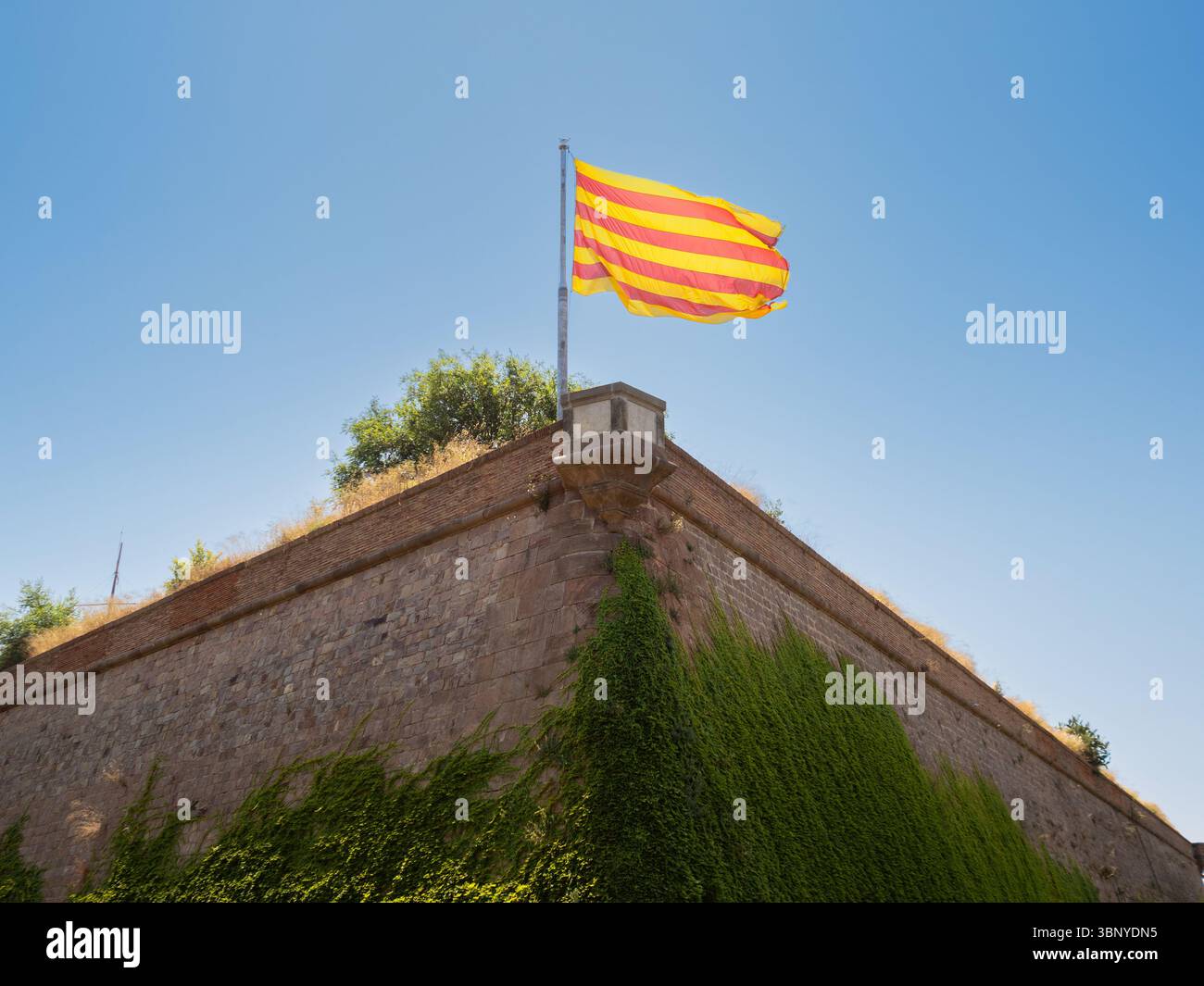 La bandiera catalana, la Senyera, che sventola orgogliosamente dalle storiche merlature in pietra del Castello di Montjuic a Barcellona, in Spagna, in una giornata limpida. Foto Stock