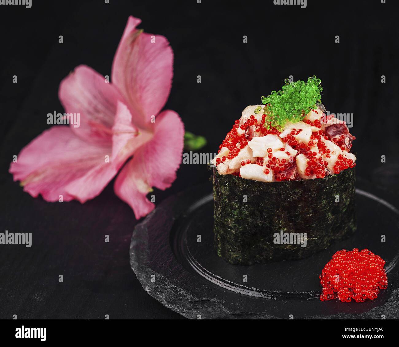 Cibo natura morta, sushi Gunkan maki ricoperto di tobiko e caviale rosso è presentato su un piatto di ardesia, accompagnato da wasabi e un fiore rosa Foto Stock