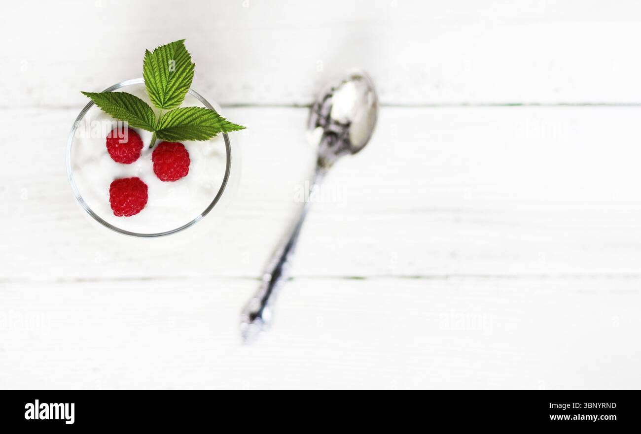Un bicchiere con yogurt al latte, lamponi maturi e una foglia verde su un tavolo di legno bianco con una vista dall'alto dello spazio copia, fuoco selettivo Foto Stock