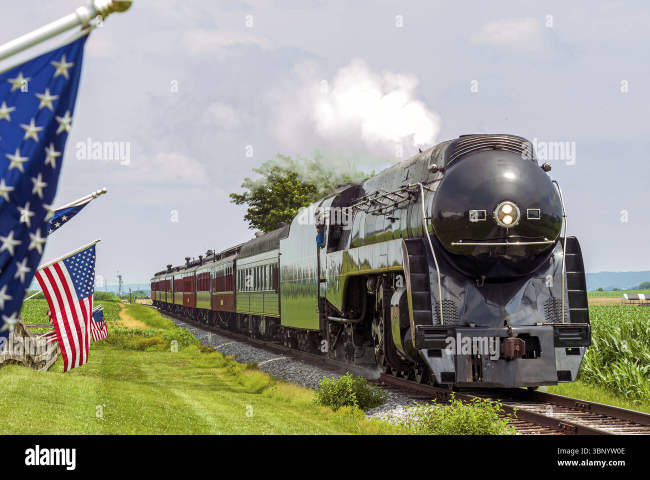 Un treno passeggeri a vapore restaurato si avvicina come American Flags Wave on a Fence at the Train Travelling Thru Farmlands in un giorno estivo soleggiato Foto Stock