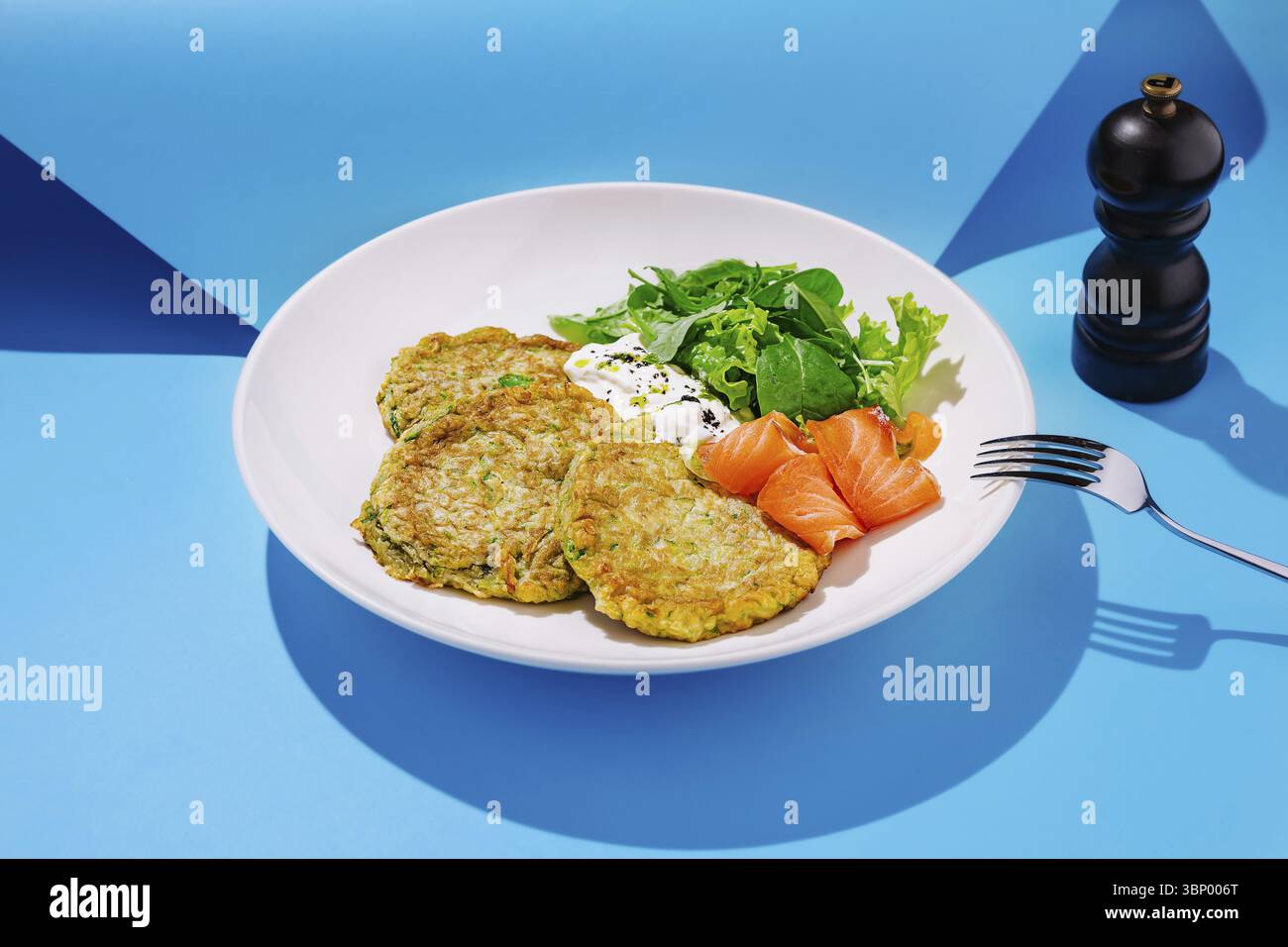 Food STILL-Life, Un piatto di frittelle di zucchine viene presentato insieme al salmone affumicato e a un'insalata fresca di verdure a foglia verde. Lo sfondo blu brillante aggiunge un Foto Stock