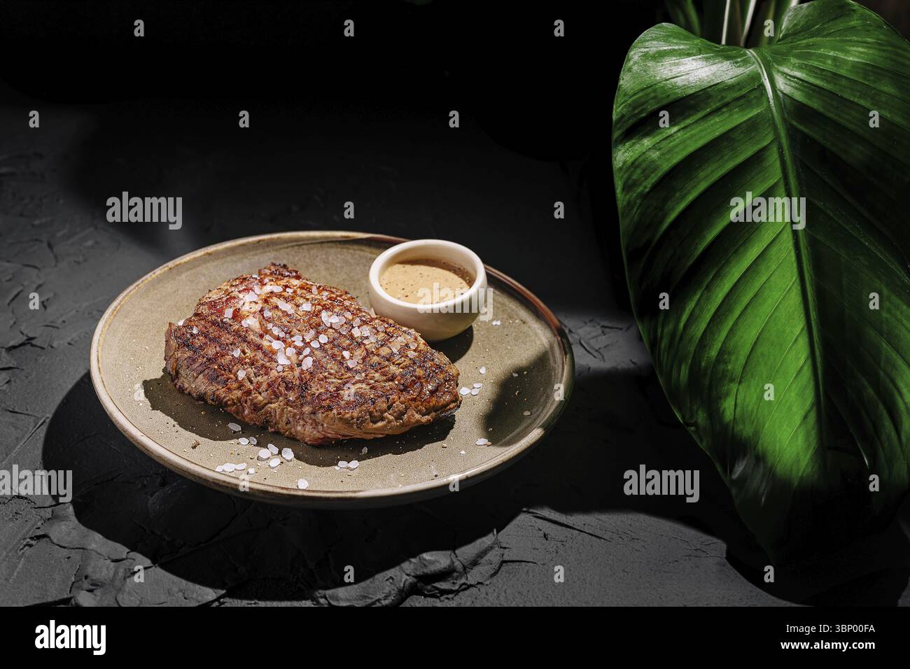 Cibo natura morta, succosa bistecca cucinata alla perfezione è posta su un piatto testurizzato, accompagnato da una piccola ciotola di salsa da immersione. Viene aggiunta una grande foglia verde Foto Stock