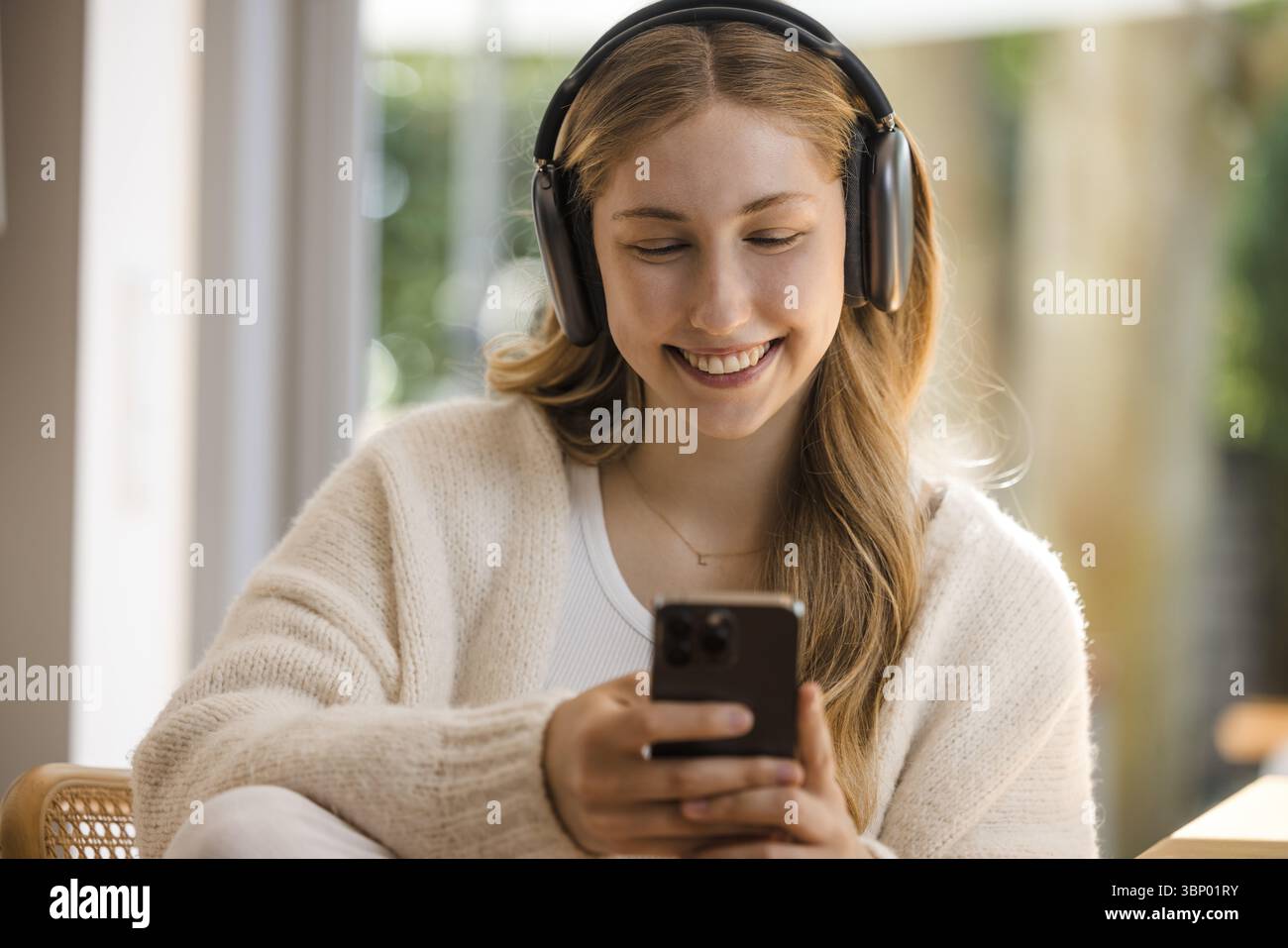 Giovane donna bionda con la parte superiore bianca si siede rilassata al tavolo con le cuffie e il telefono cellulare, sorridendo Foto Stock