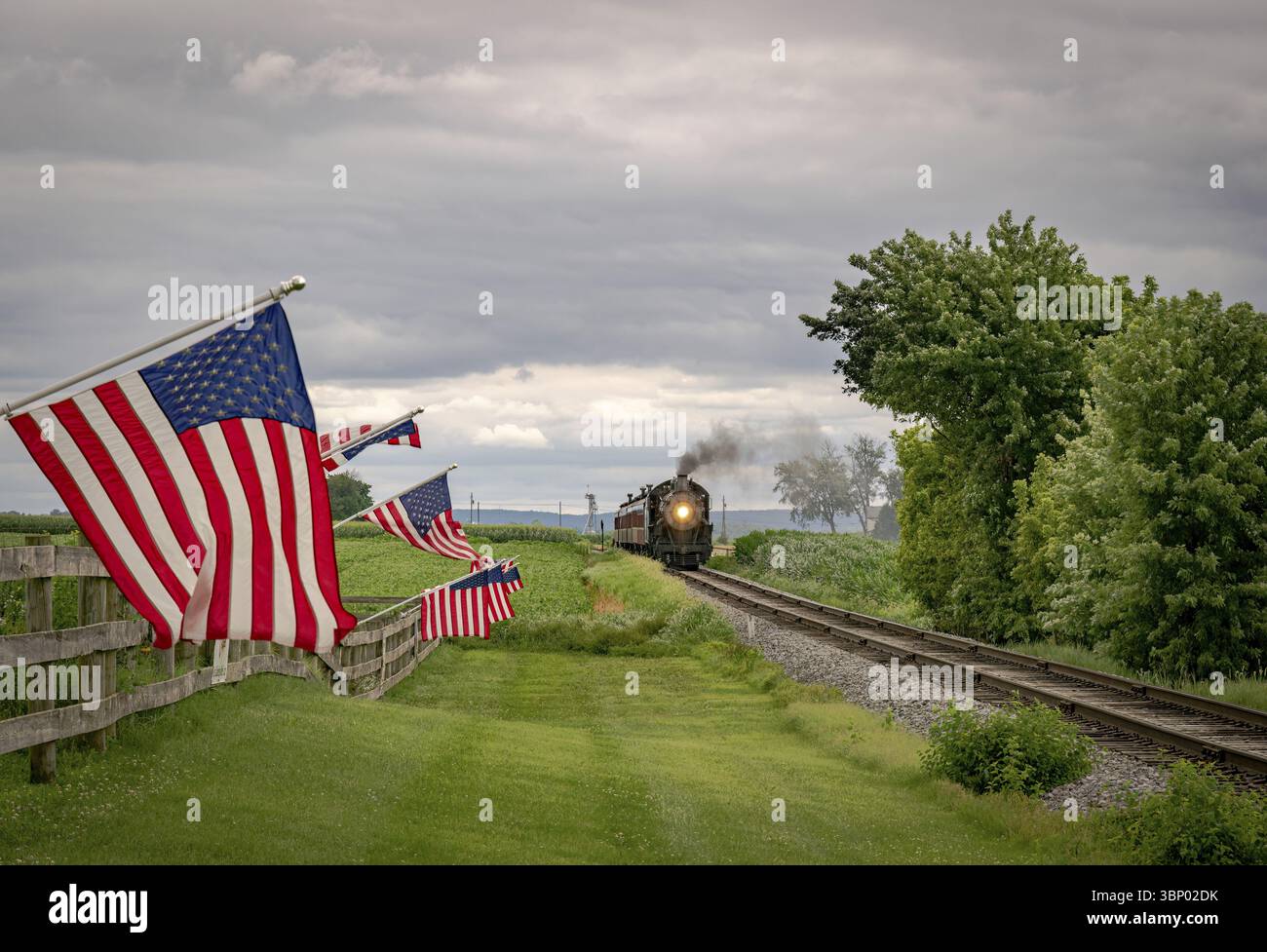 In un giorno nuvoloso le bandiere americane sventolano nel vento, su una recinzione, mentre un treno a vapore passeggeri si avvicina Foto Stock