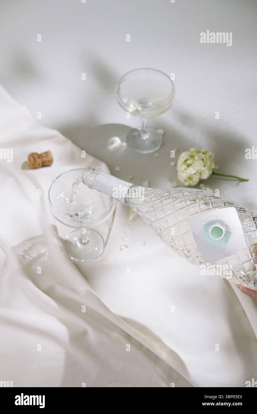 Food STILL-Life, Una mano versa una bevanda chiara da un'elegante bottiglia di vetro in due bicchieri coupé su tessuto bianco. Un delicato fiore bianco aggiunge un Foto Stock