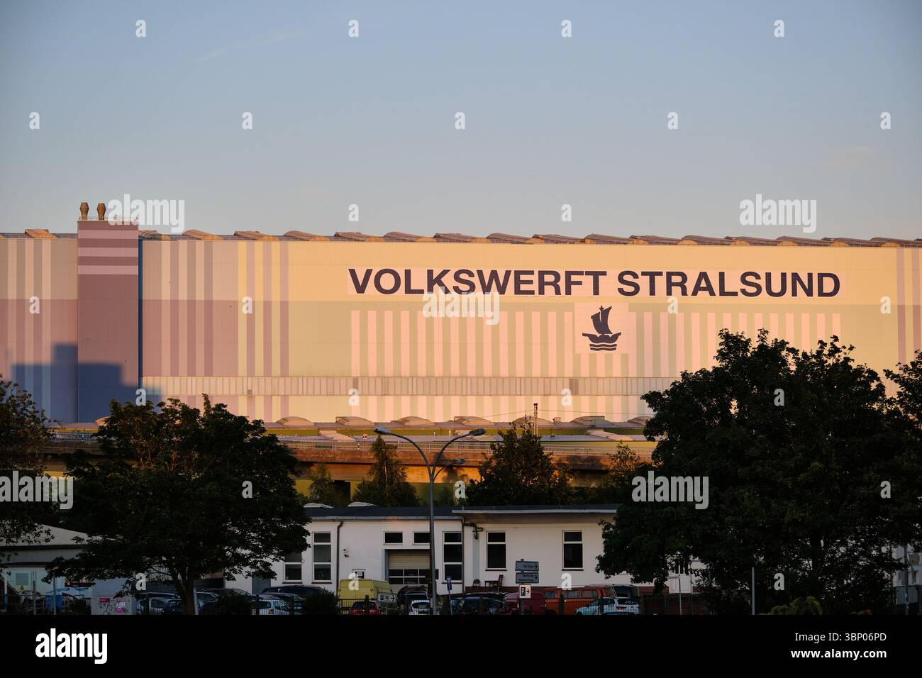 30 giugno 2025 - Stralsund-Germania: La sala del cantiere Volkswerft si illumina con il caldo sole della sera sotto un cielo azzurro limpido con primo piano industriale e verde Foto Stock