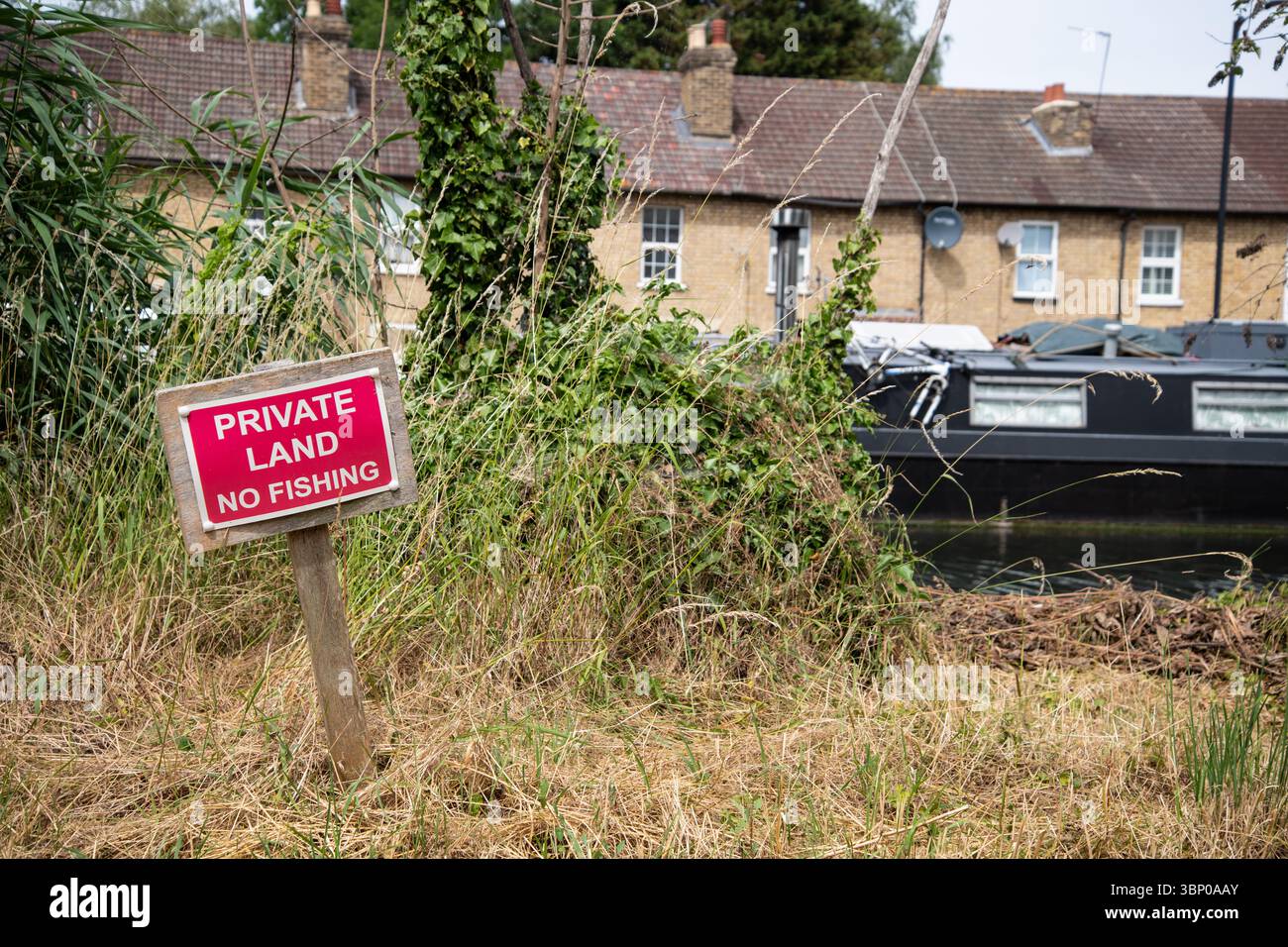 Red and white "Private Land No Fishing" sign on the bank of the River Lea Navigation at Government Row, Enfield, with a canal boat and houses behind. Foto Stock