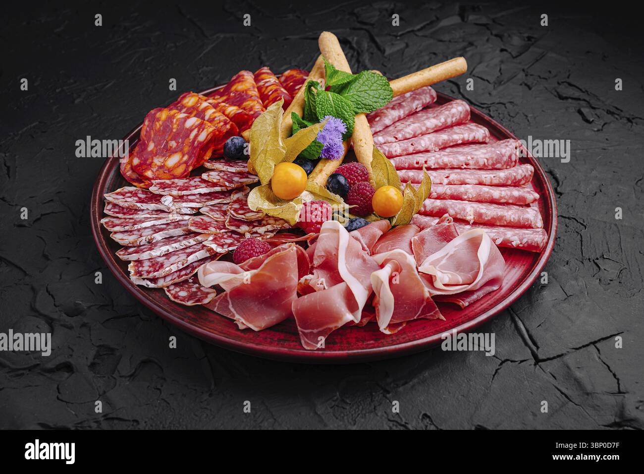 Food STILL-Life, Un vivace assortimento di salumi, tra cui salame e prosciutto, è elegantemente esposto su un piatto scuro, circondato da fru fresca Foto Stock