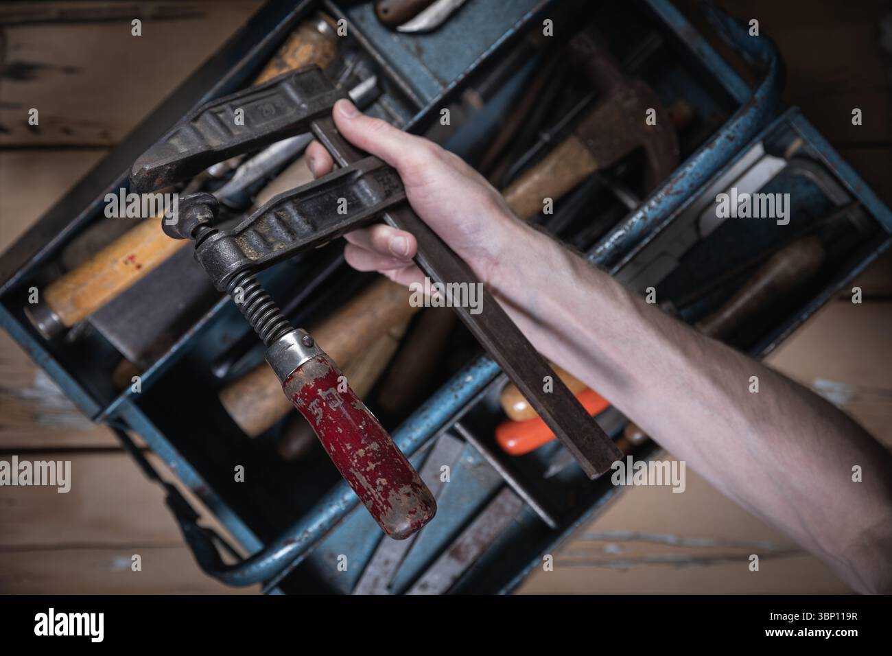 Mano che regge un vecchio morsetto con manico in legno rosso davanti a una cassetta degli attrezzi aperta in metallo blu contenente utensili vintage su un tavolo in legno usurato. Foto Stock