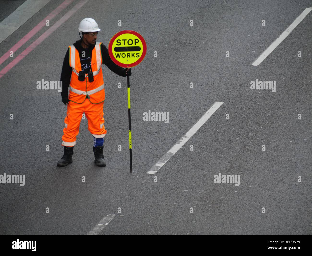 Un traffico edile marshall tiene un cartello "Stop Works" mentre gestisce il flusso del veicolo attraverso una strada trafficata della City di Londra, Regno Unito. Essenziale per mantenere la sicurezza stradale durante i progetti di sviluppo urbano in corso Foto Stock
