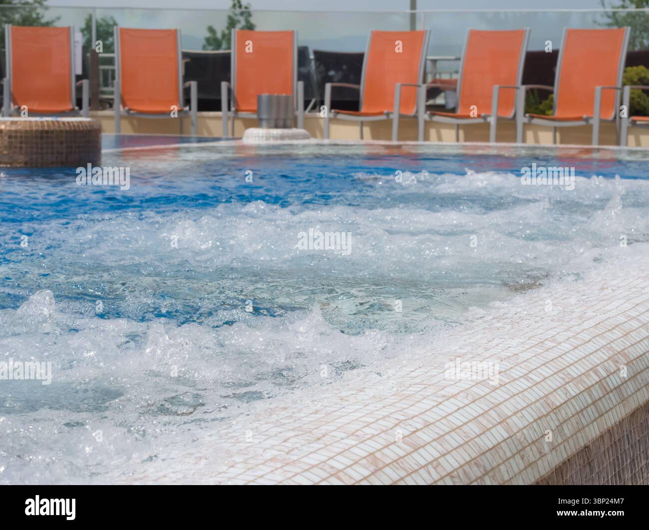 Vista ravvicinata di una gorgogliante vasca idromassaggio con piastrelle a mosaico bianche e marroni, con sedie a sdraio arancioni sfocate disposte sullo sfondo sotto Clear Foto Stock