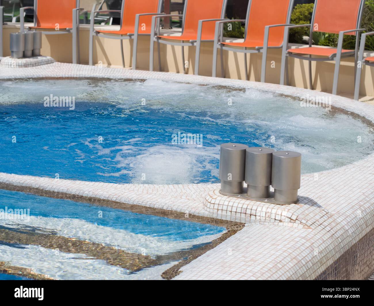 Una vista ravvicinata di una jacuzzi del resort con piastrelle a mosaico bianche e marroni, che mostra la sua invitante acqua frizzante blu e le vivaci sedie a sdraio arancioni Foto Stock