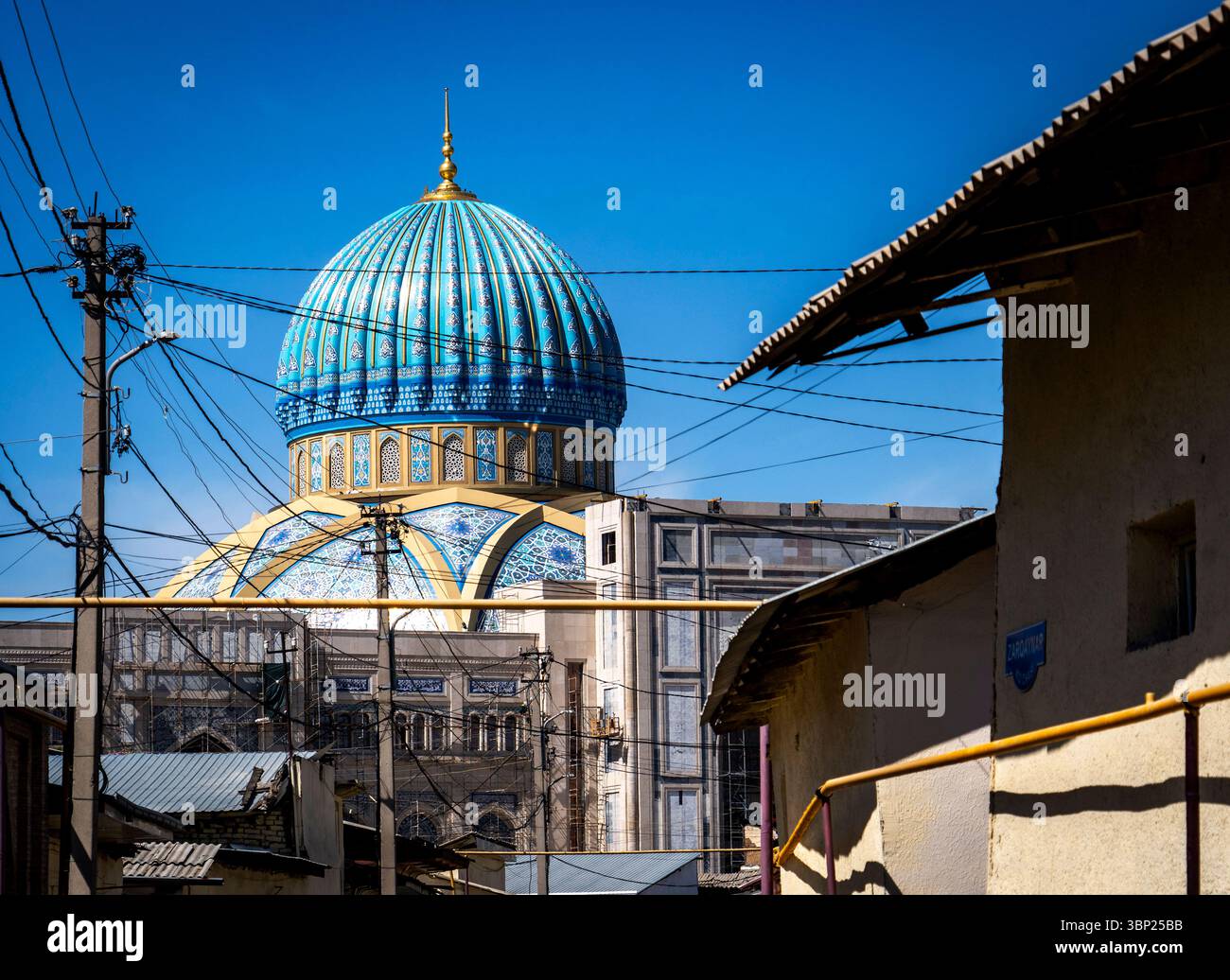 Una vivace cupola blu di una moschea si erge sopra edifici urbani, incorniciata da linee elettriche contro un cielo azzurro. Foto Stock