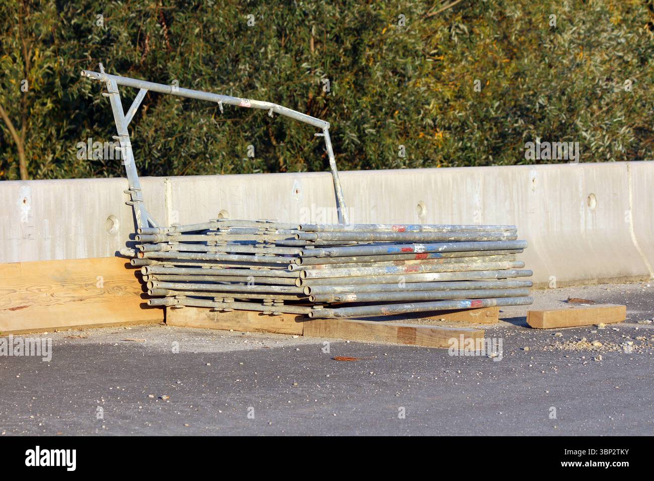 Una pila ordinata di tubi per impalcature in metallo galvanizzato poggia su tavole di legno accanto a una barriera in calcestruzzo in un cantiere stradale, con alberi Foto Stock