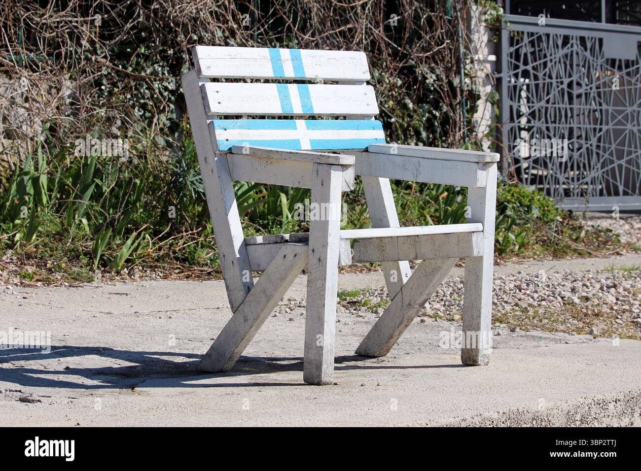 Una sedia rustica in legno bianco con audaci strisce blu si erge da sola su un sentiero di cemento illuminato dal sole, la sua vernice invecchiata e la struttura robusta evocano un senso di esterno Foto Stock