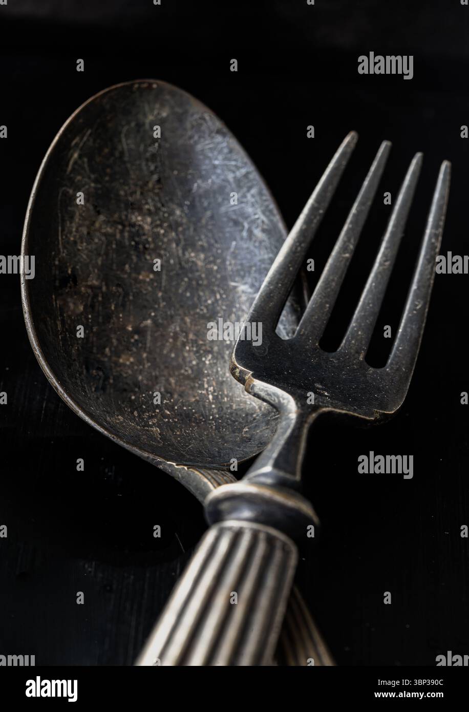 Antiche posate vintage su sfondo nero. Cucchiaio e forchetta in metallo nero incrociato in tonalità scura, che ne esalta la consistenza e il design, usali come Foto Stock