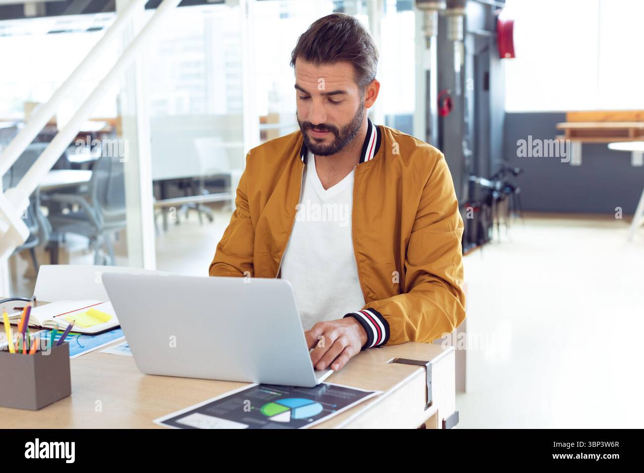 Uomo che indossa un bomber che lavora alla scrivania condivisa utilizzando un computer portatile e visualizzando le carte stampate Foto Stock