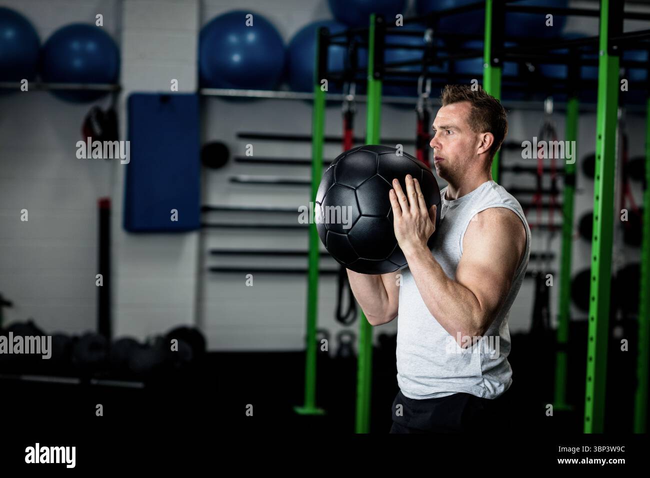 Uomo di mezza età che solleva palla medica nera nella zona di allenamento della palestra con scaffale per squat e spazio per fotocopie Foto Stock
