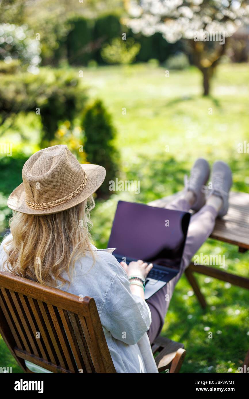 Donna che usa un notebook sotto un albero in fiore nel giardino di primavera. Concetto di lavoro freelance e lavoro in remoto Foto Stock