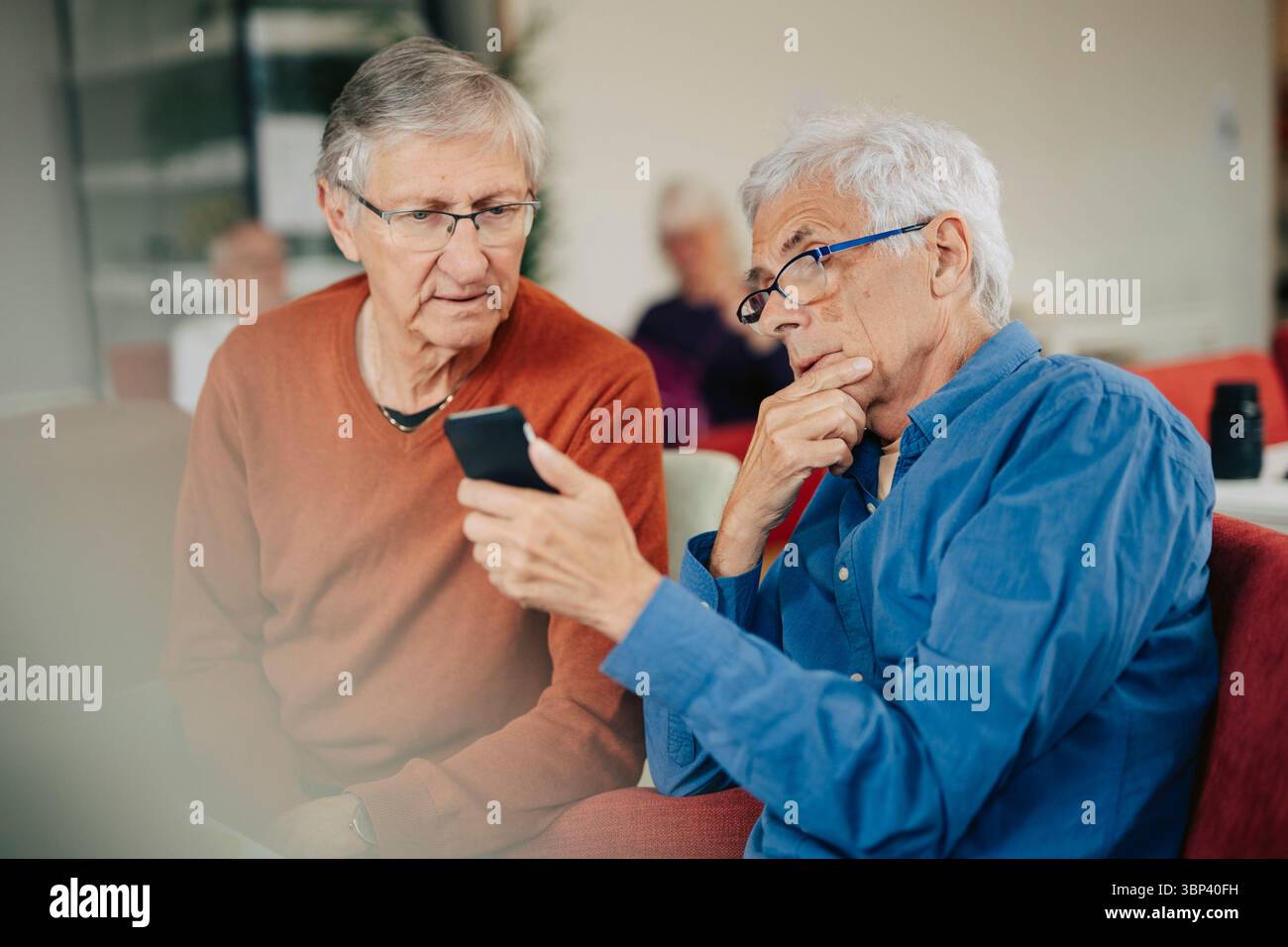 Anziani in pensione che condividono lo smartphone con un amico mentre sono seduti a casa di riposo Foto Stock