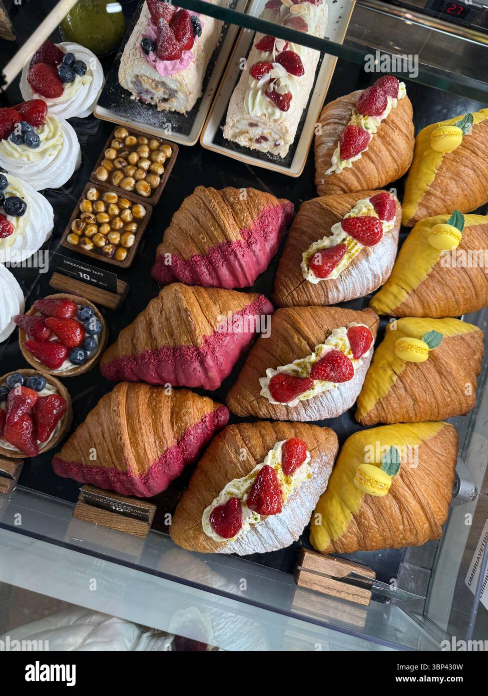 Questa foto mostra una vetrina di panetteria contenente croissant con glassa rosa e gialla, croissant ricoperti di panna e fragole, pavlovas con Foto Stock