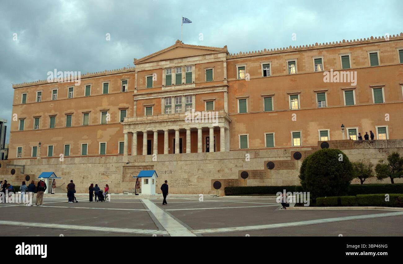 Una fotografia dell'edificio del Parlamento ellenico ad Atene, in Grecia, la legislatura unicamerale della Grecia, situata nel Vecchio Palazzo reale, affacciata su Piazza Syntagma ad Atene. L'edificio neoclassico presenta una facciata simmetrica con un portico centrale sostenuto da colonne. La bandiera greca vola dal tetto. In primo piano, le persone sono visibili nella piazza, e sono presenti anche due scatole di sentinelle, tipicamente utilizzate dalle guardie di Evzones. c. 2023. Foto Stock