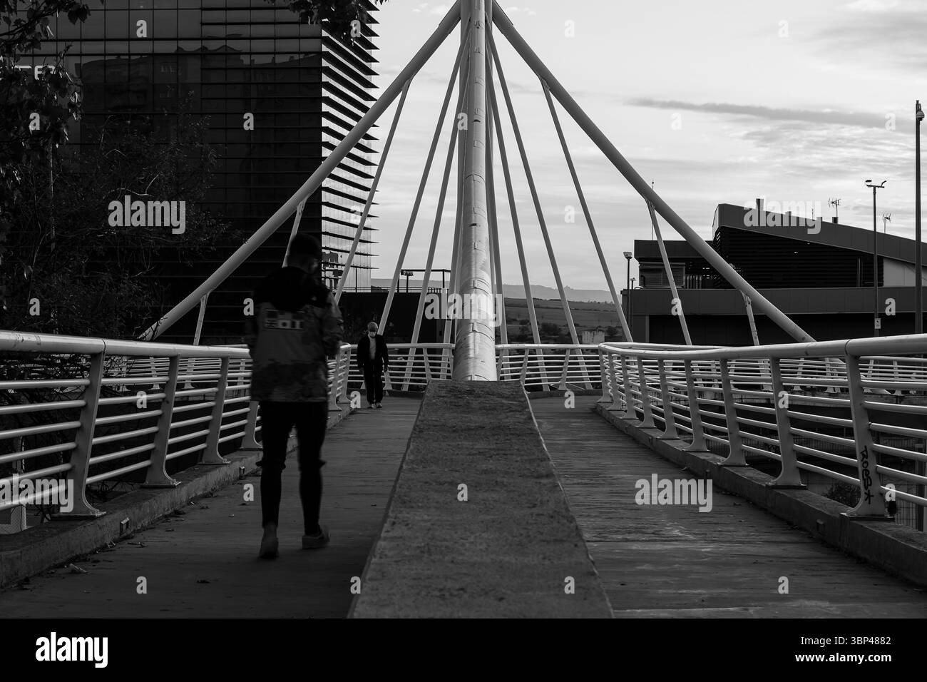 Due persone camminano su un sentiero di un ponte moderno mentre il sole tramonta, circondato da un'architettura contemporanea. Foto Stock