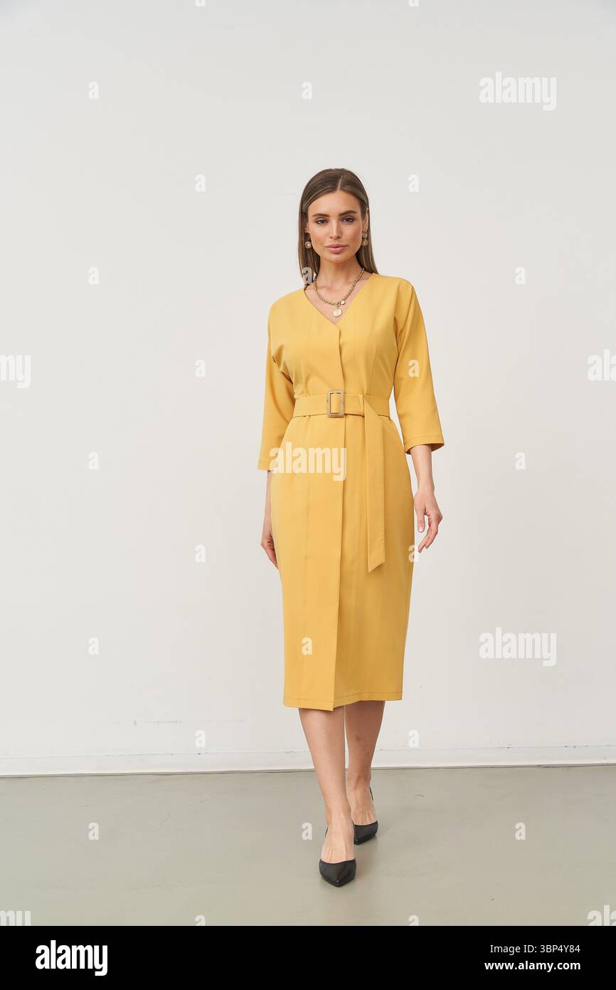 Abito al ginocchio giallo con cintura indossato da una donna con lunghi capelli marroni. Indossa scarpe nere e collane a strati, in piedi con sicurezza, con le mani rilassate Foto Stock