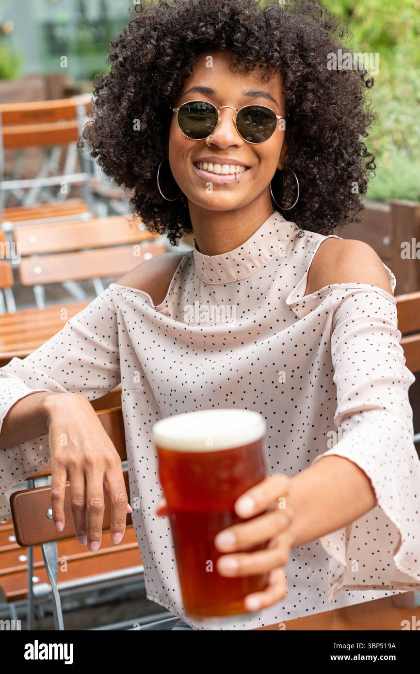 Giovane donna nera con occhiali da sole che tiene una pinta di birra e sorride in un pub all'aperto Foto Stock